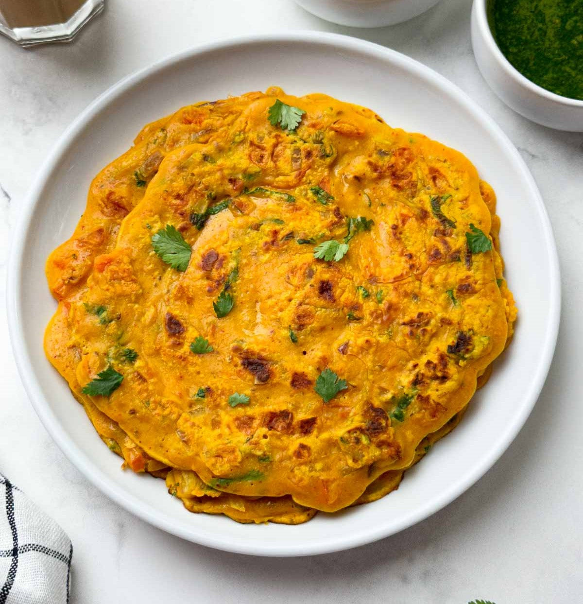 Stack of besan chilla on a white plate with green herbs, on a light gray surface.